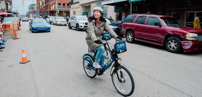 Young woman riding a bicycle with the POGOH logo on the front. She is riding down a busy city street surrounded by shops and other cars driving down the road.