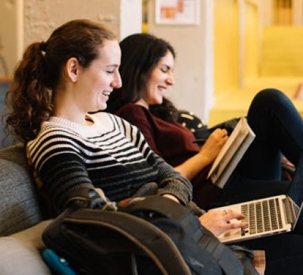 Female students sitting and smiling working on laptop and reading a book